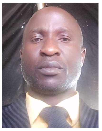 Pastor Olayinka Olatunji wearing a yellow shirt, a dark blue tie, and a dark striped suit jacket, standing against a dark background.